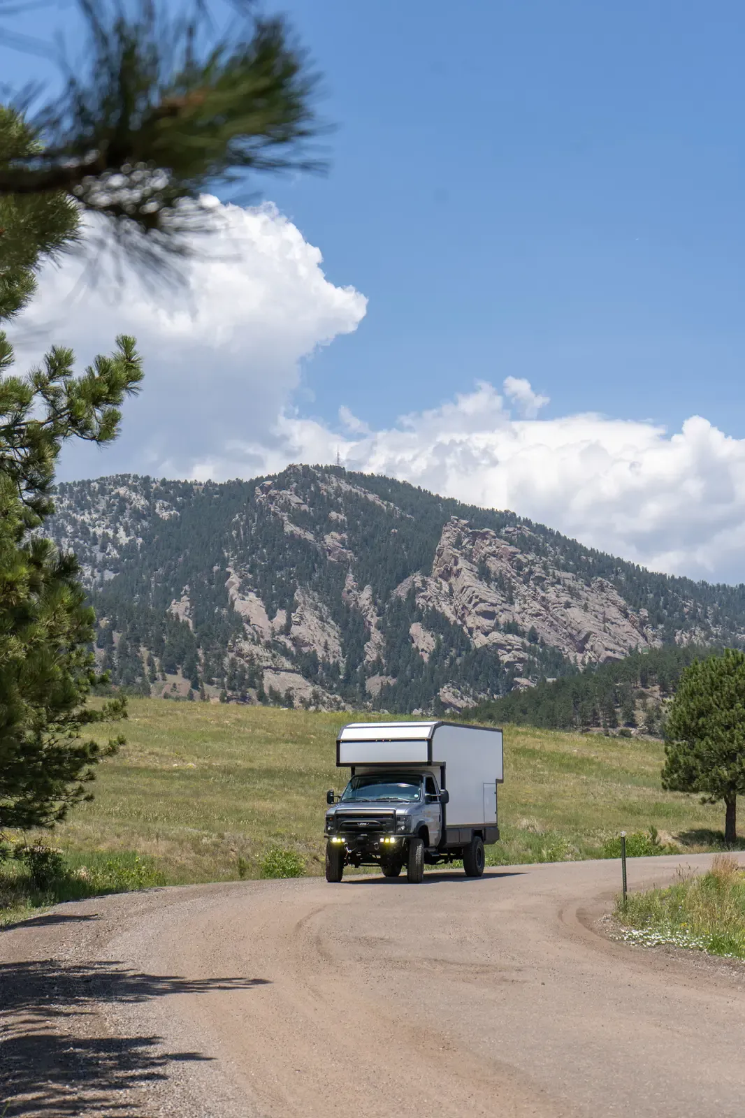 Silver-cab Ford E-450 expedition camper approaching on a dirt road with the foothills behind