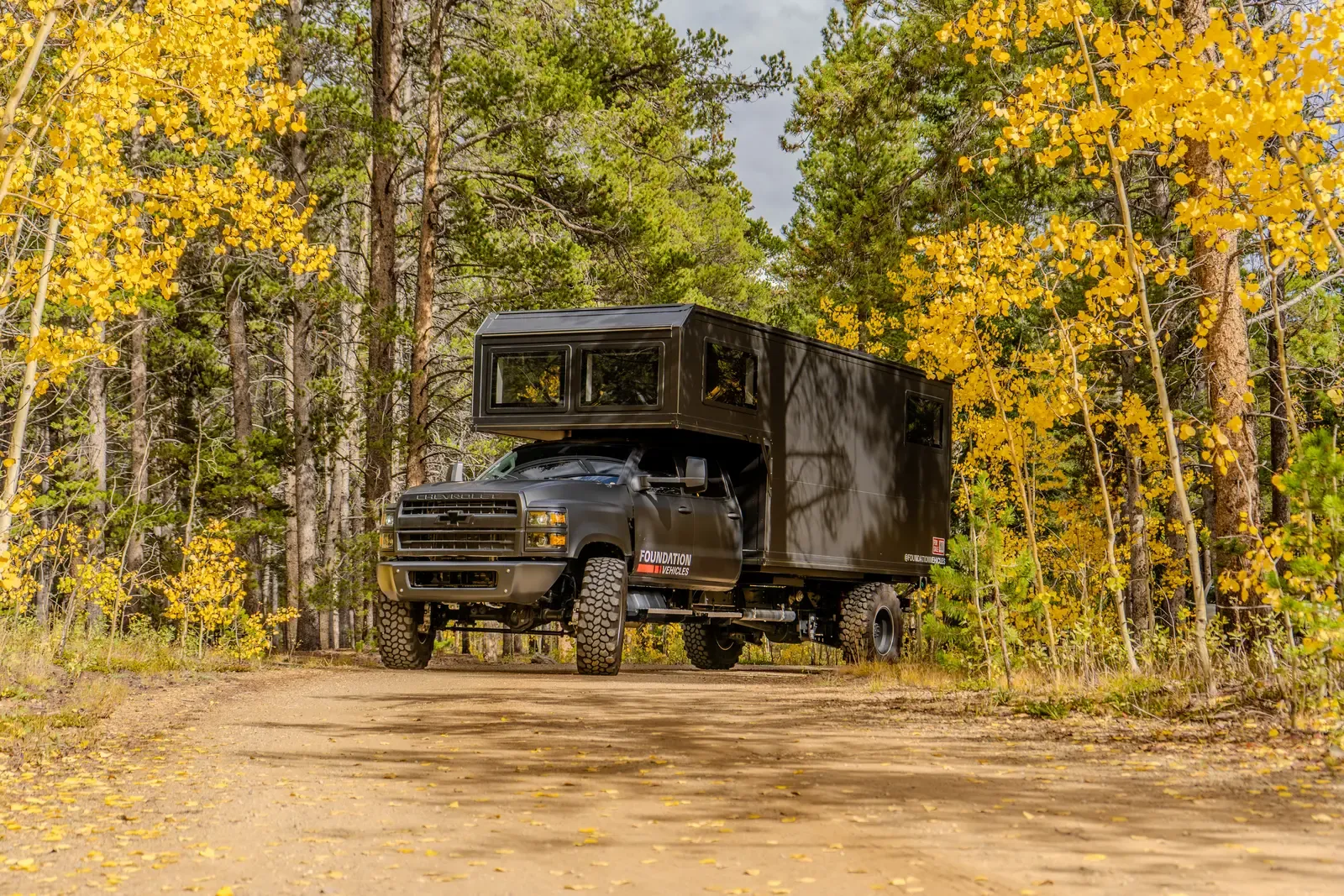 Blacked-out Chevrolet 6500 expedition camper, side profile driving through a stand of golden aspens