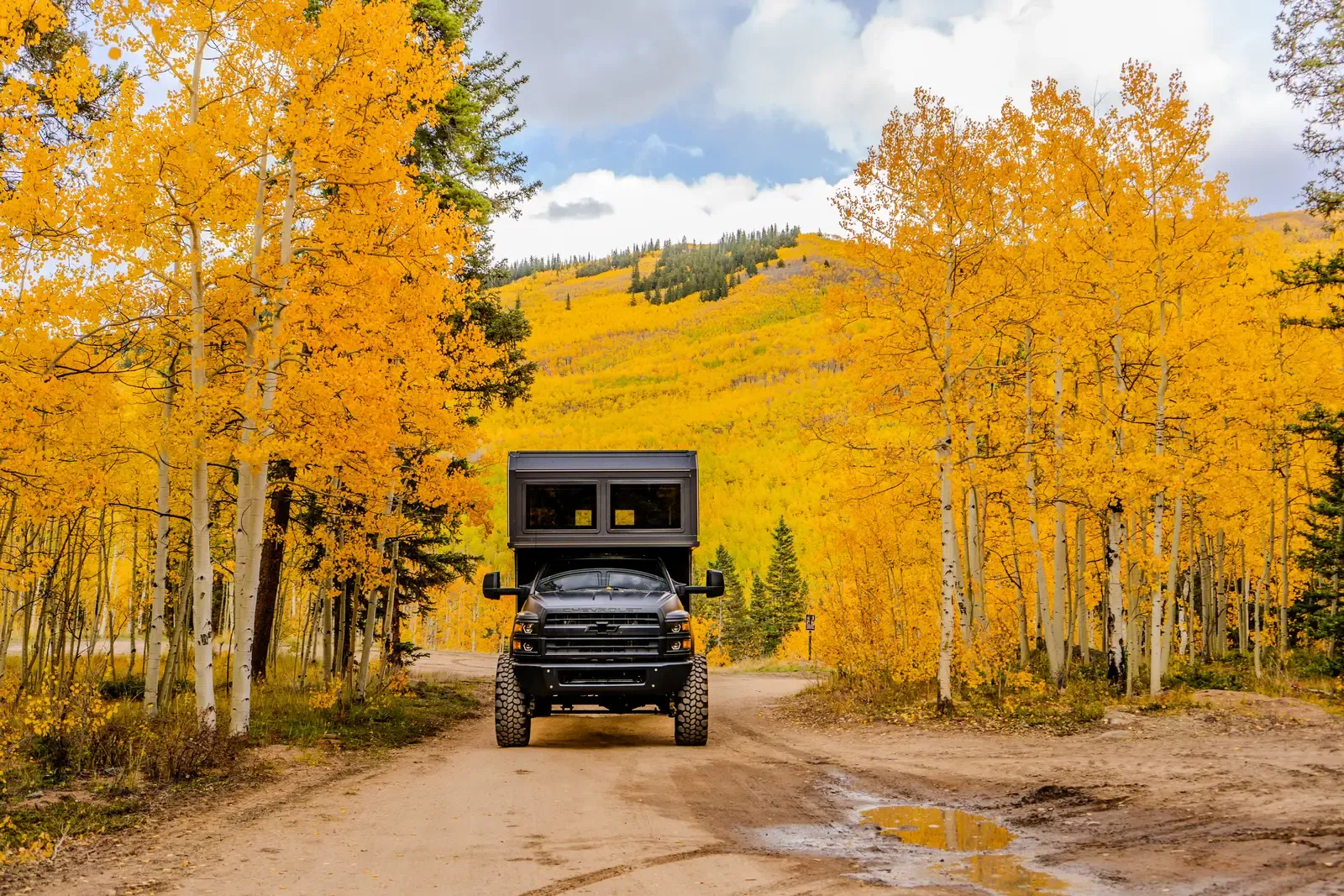 Blacked-out Chevrolet 6500 4x4 expedition camper, front three-quarter view on a mountain dirt road in autumn
