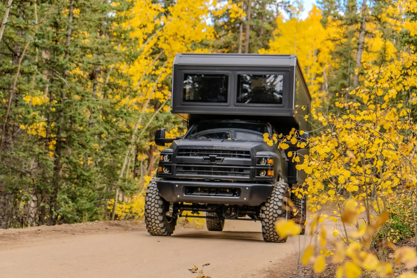 Blacked-out Chevrolet 6500 expedition camper, side view on a dirt road through an autumn aspen grove