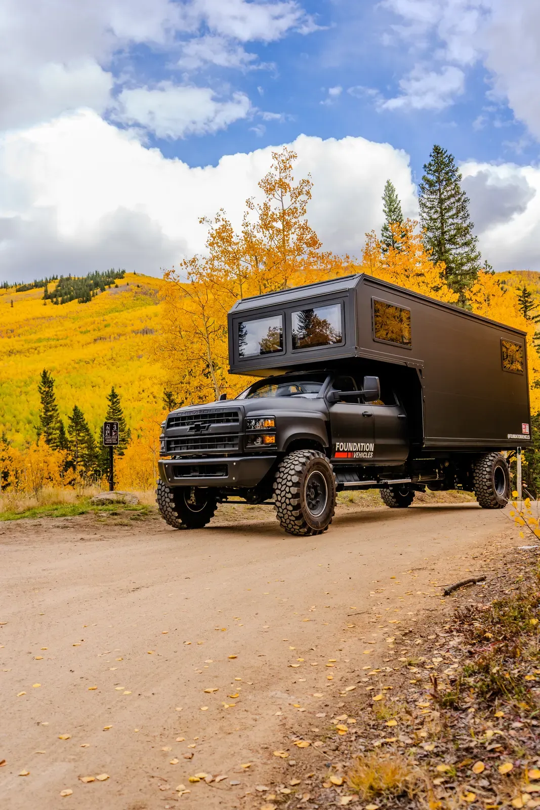 Blacked-out Chevrolet 6500 4x4 expedition camper, front three-quarter view amid bright yellow aspens