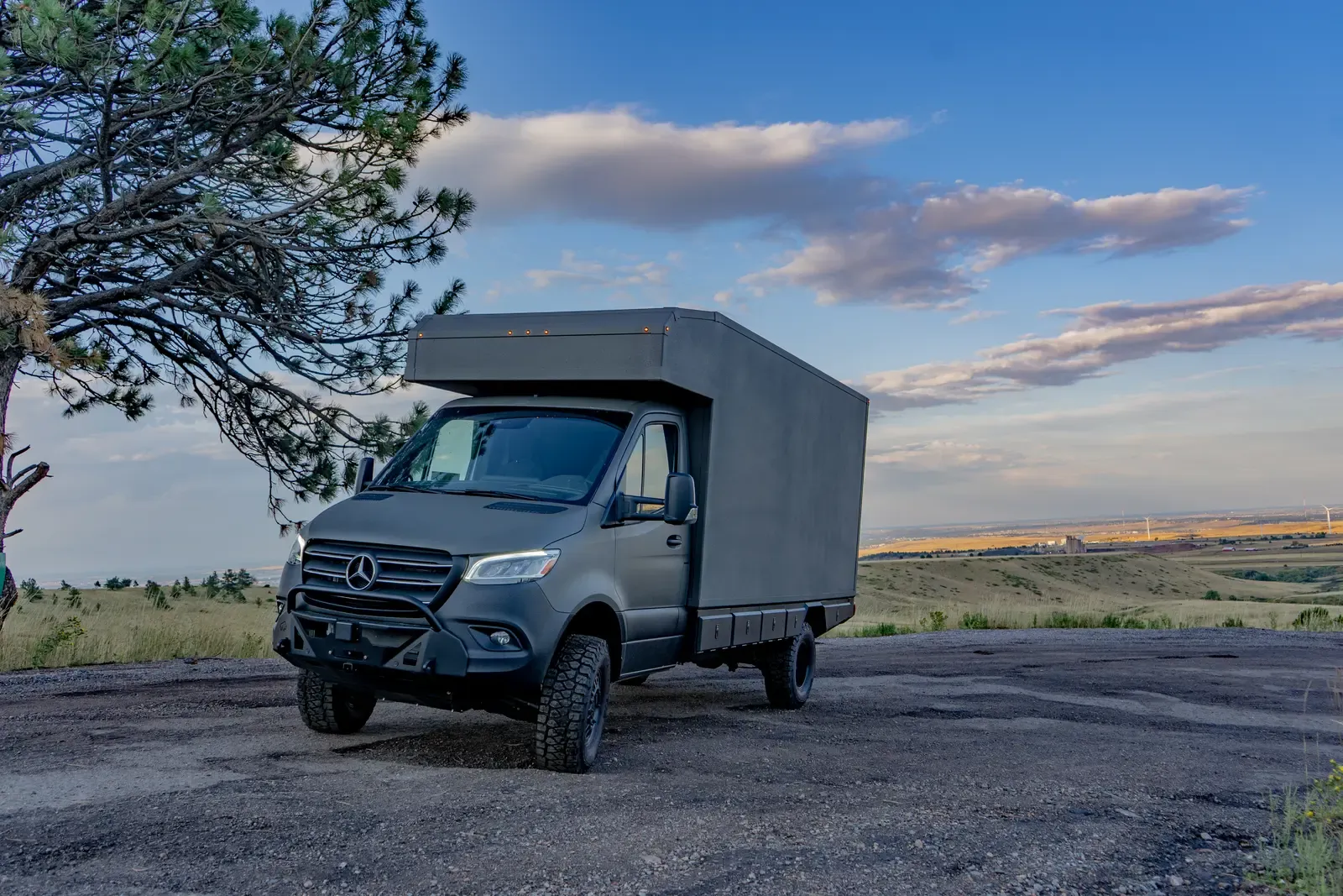 Matte-olive FV-170 demo composite camper on a Mercedes-Benz Sprinter 170, front three-quarter view at dusk beside a pine