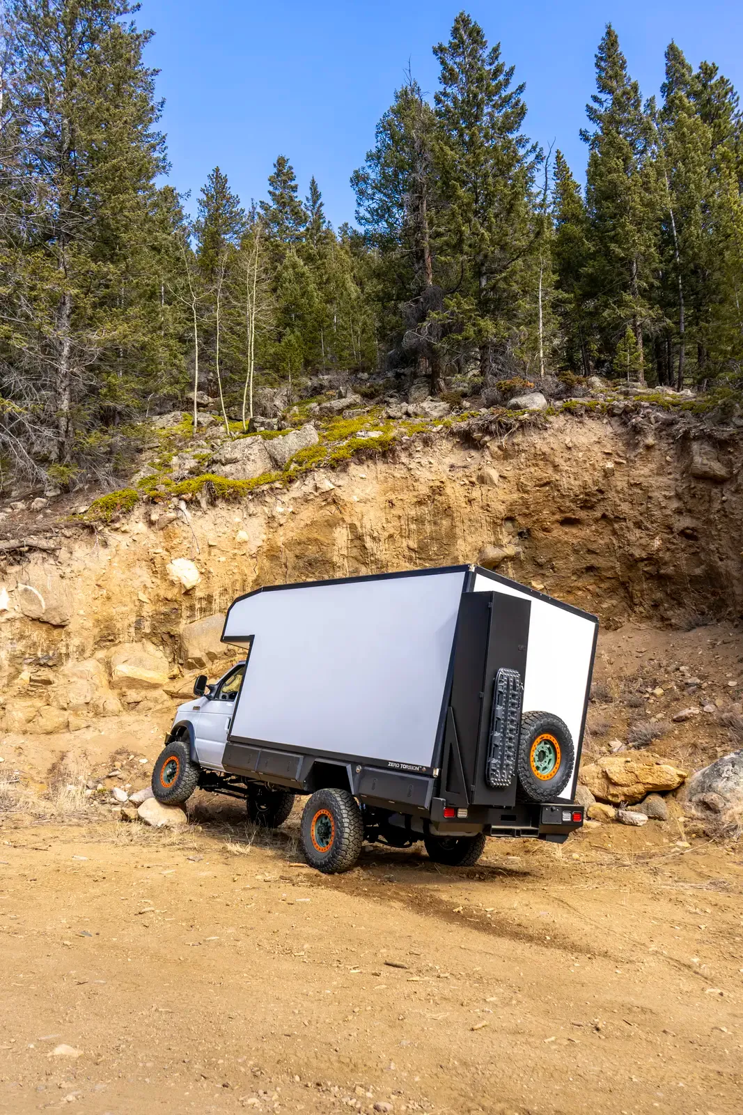 White E-158 composite box camper on a Ford E-Series 4x4 climbing a steep rocky embankment, rear three-quarter view