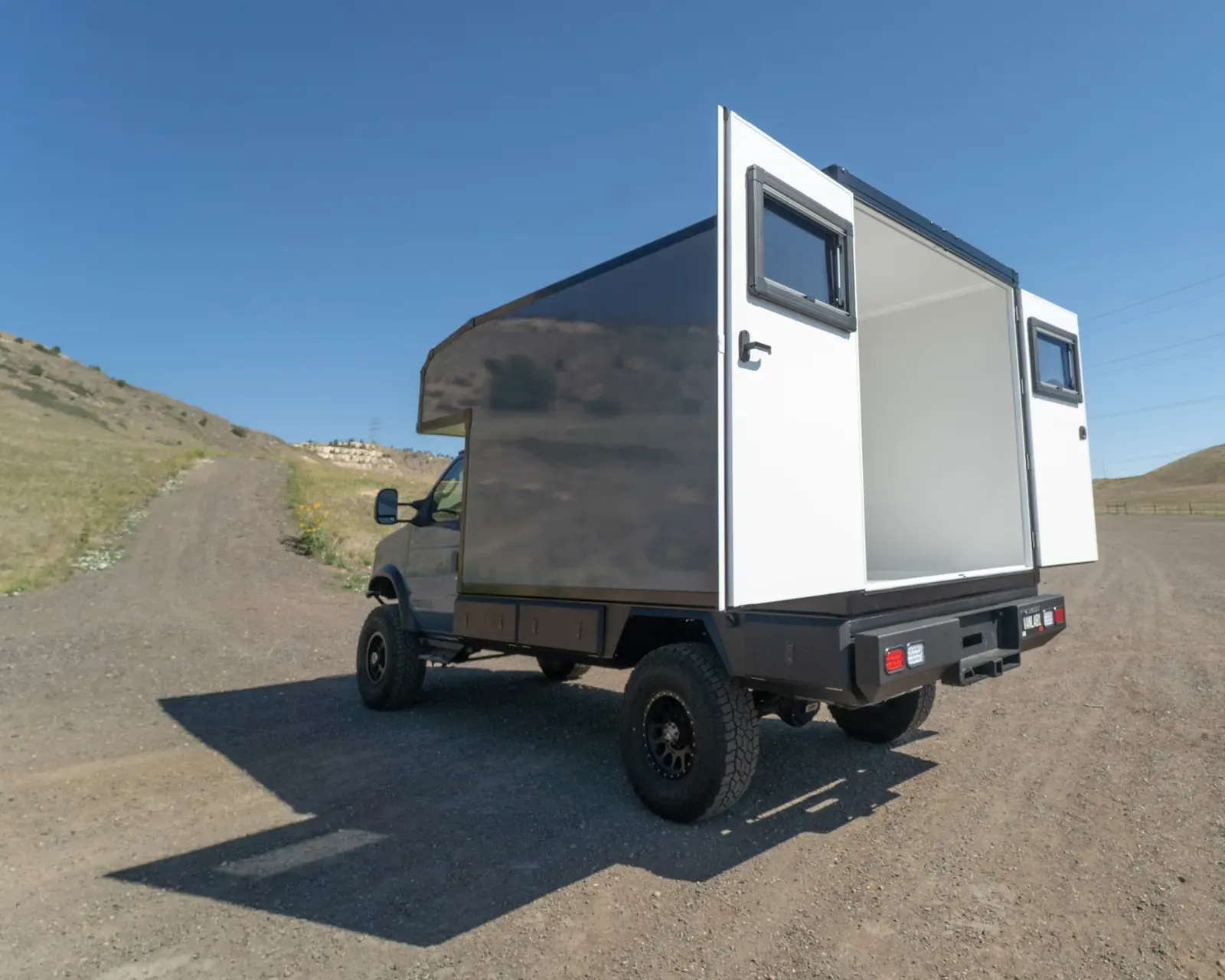 Cavalry Blue and white Ford E-Series E-158S 4x4 composite camper, rear three-quarter view on a dirt road