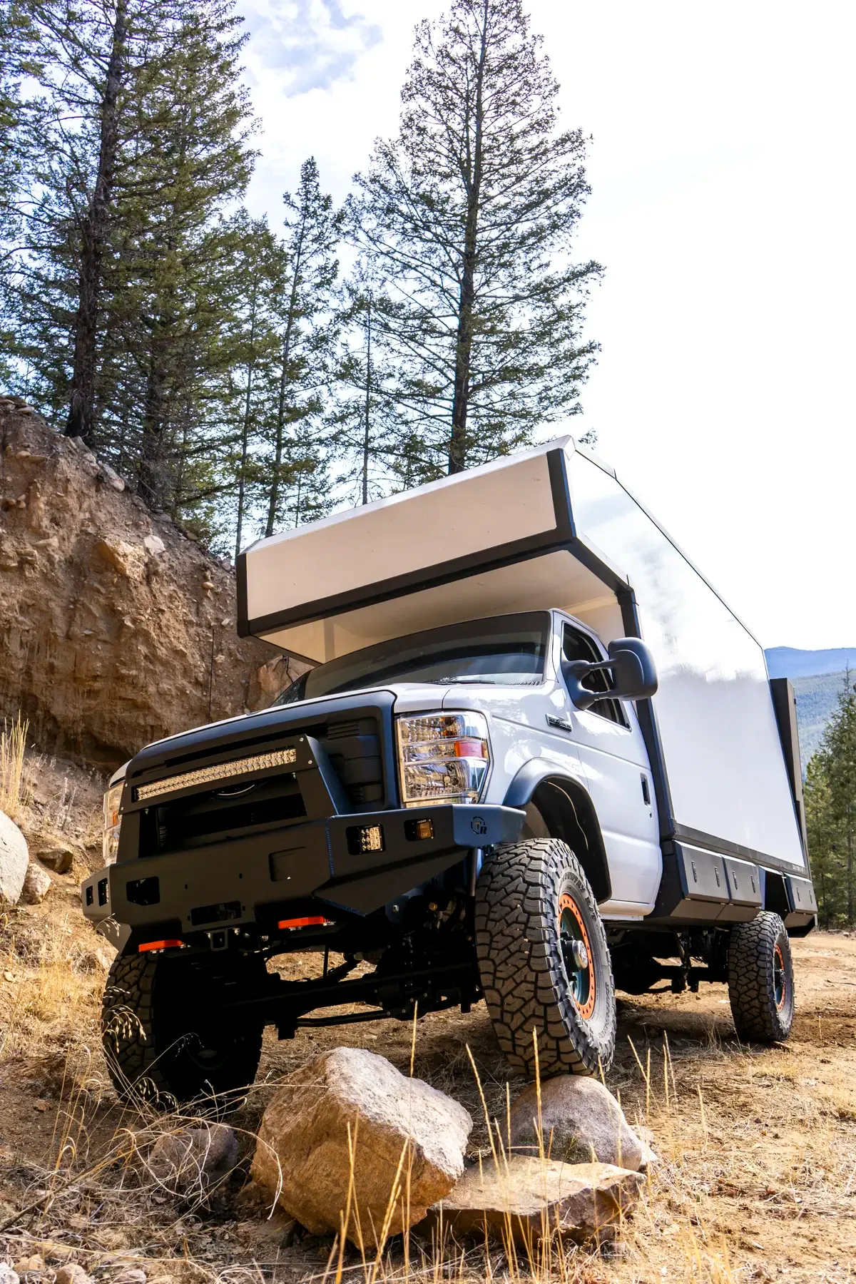 Ford E-Series 4x4 composite camper on a rocky off-road trail, low front three-quarter view showing the lifted suspension, off-road bumper and all-terrain tires