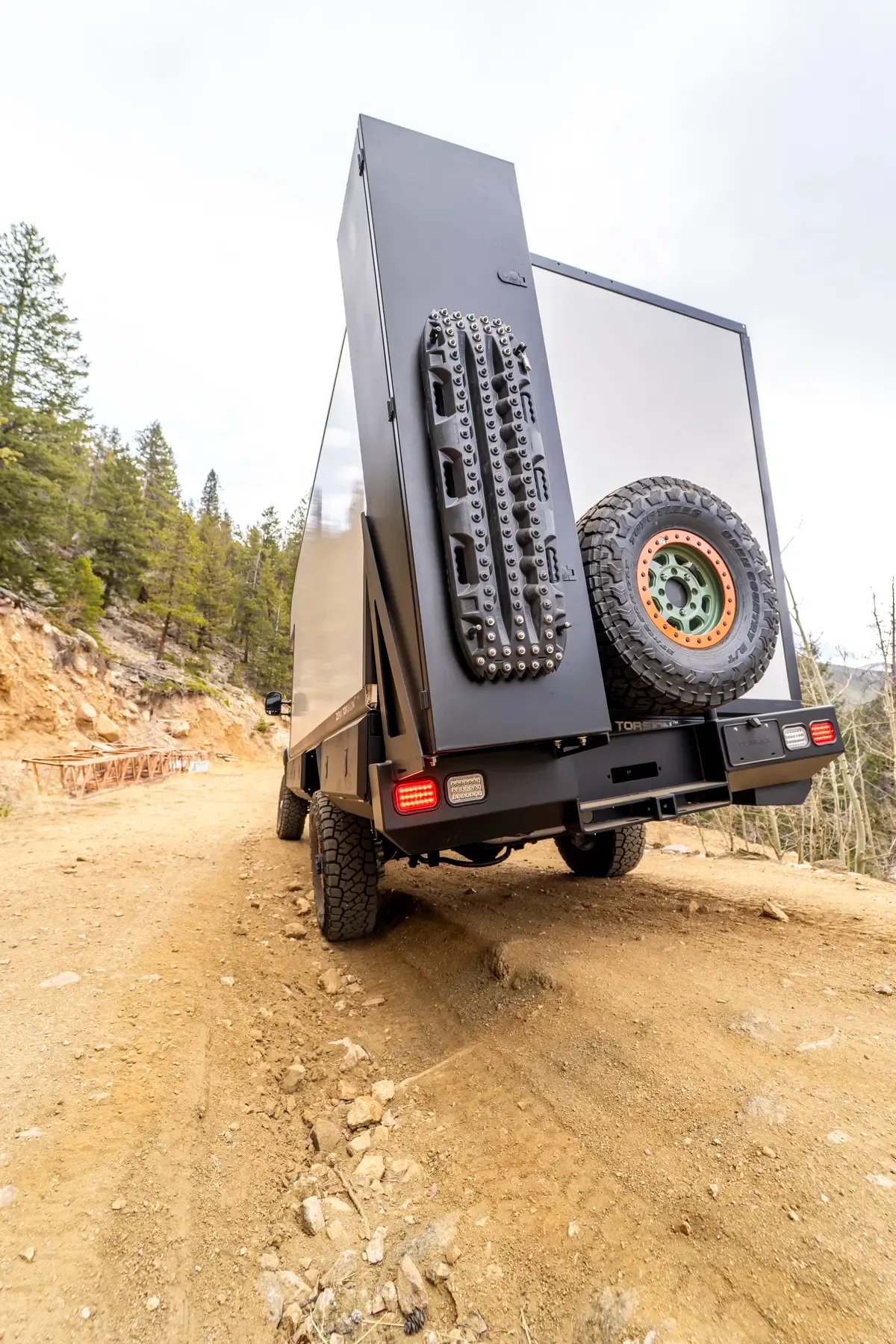 Rear view of a Foundation Vehicles E-Series composite camper on an uneven rocky trail, with traction boards and a spare all-terrain tire mounted on the rear door