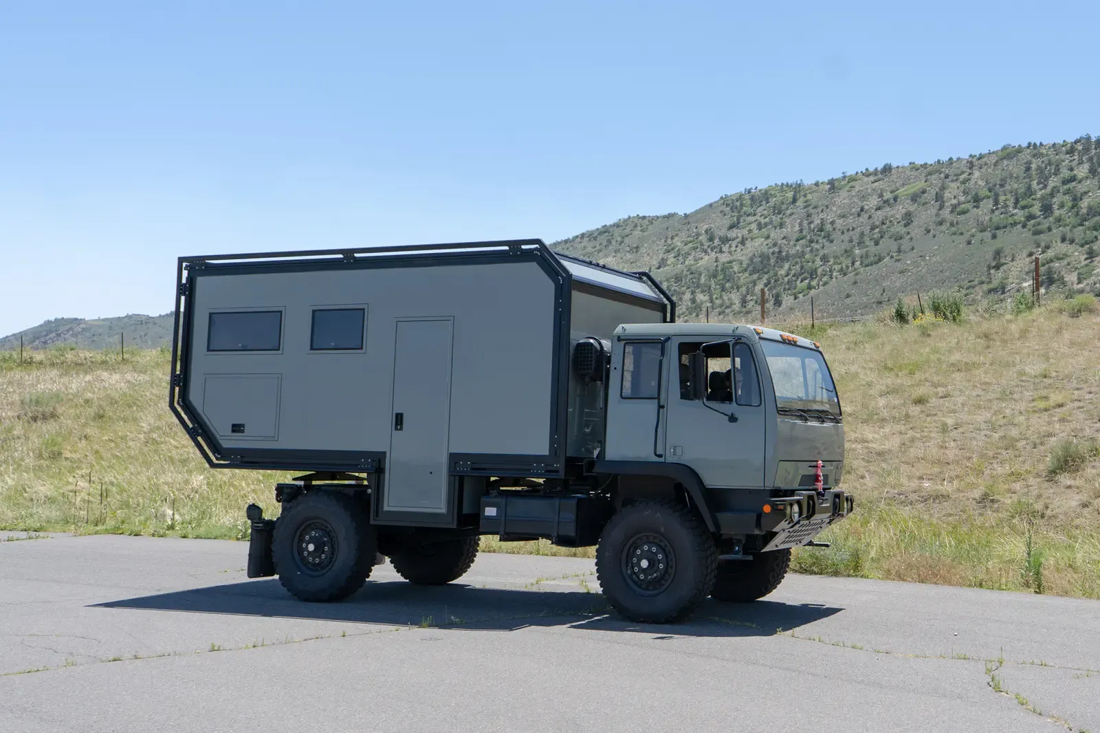 Sage-green LMTV expedition truck with a composite camper body on a paved road, side-rear view with mountains