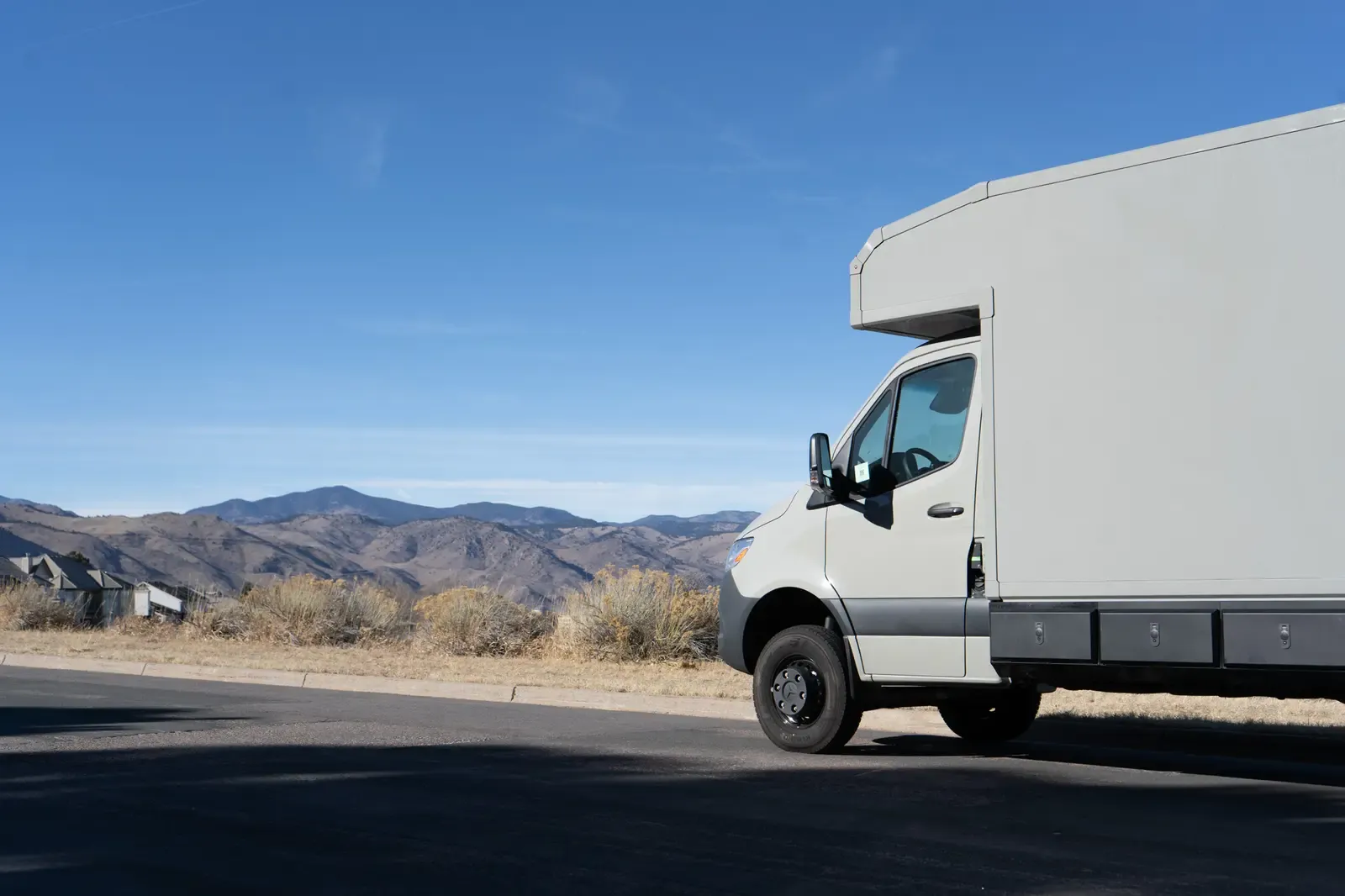 White commercial box body on a Mercedes-Benz Sprinter cab-chassis, front three-quarter view on a road with mountains