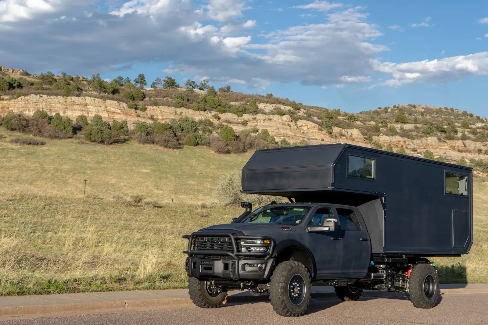 Black composite flatbed camper on a Ram 5500 cabover, front three-quarter on a country road below foothills