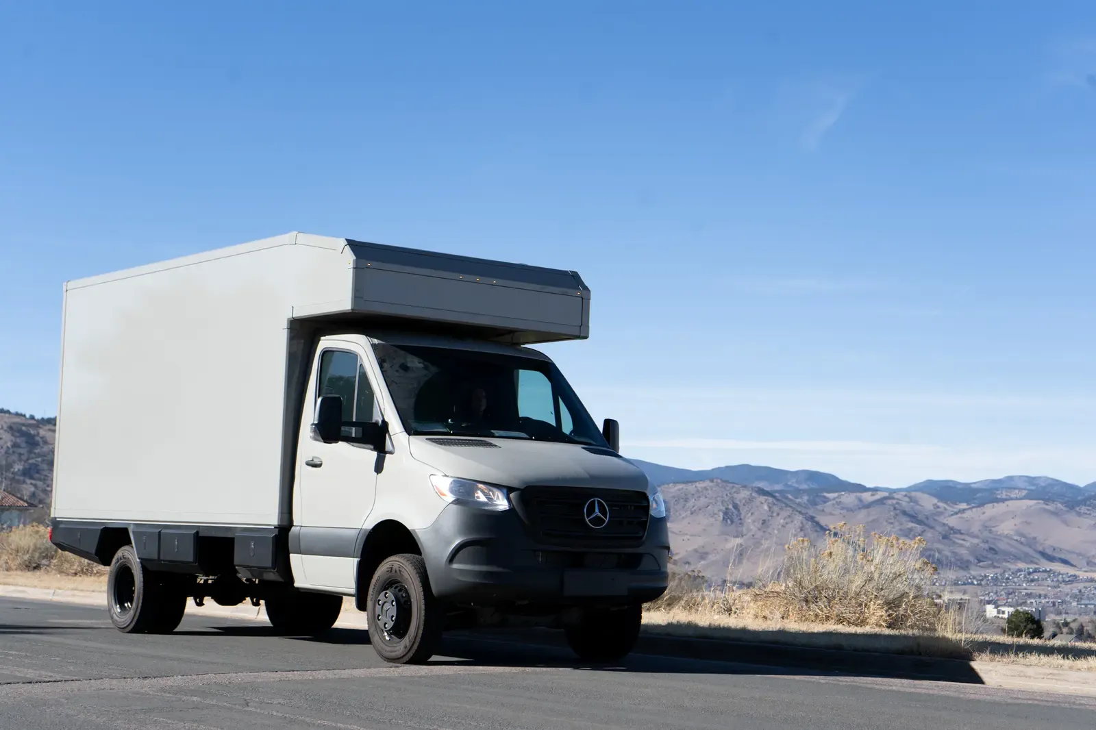 Stone grey commercial box body on a Mercedes-Benz Sprinter cab-chassis, front three-quarter view on a hilltop with mountains behind