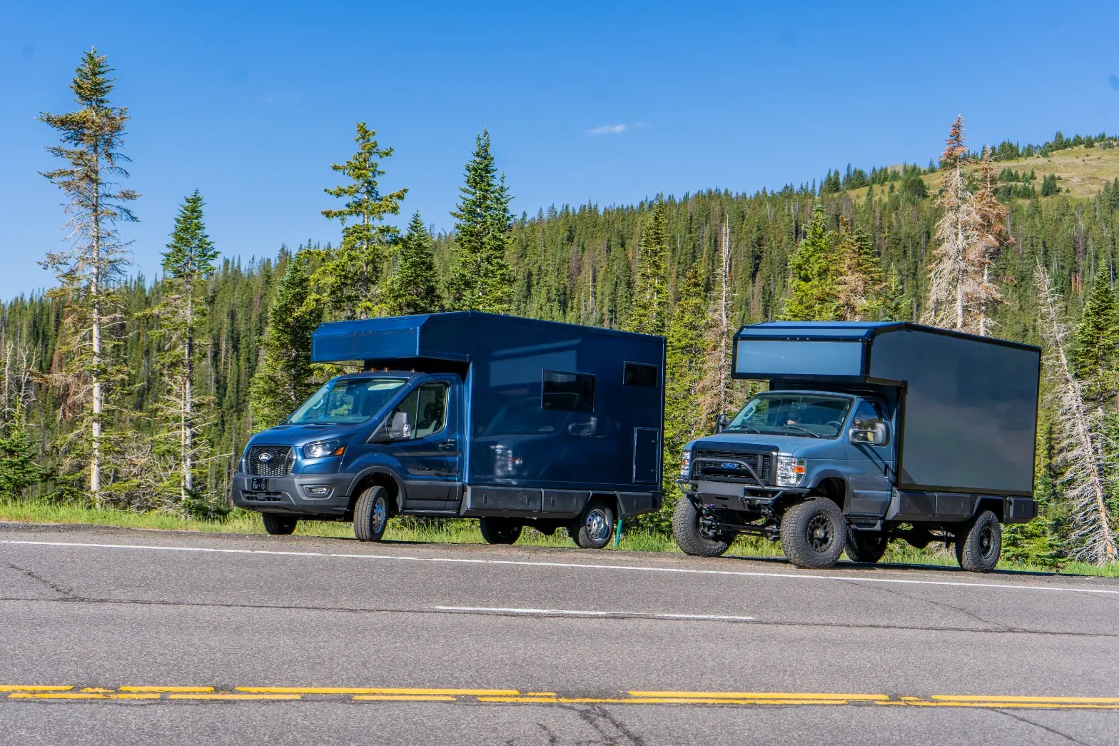 A Ford Transit composite camper and a lifted Ford E-Series composite camper parked together on a forest highway