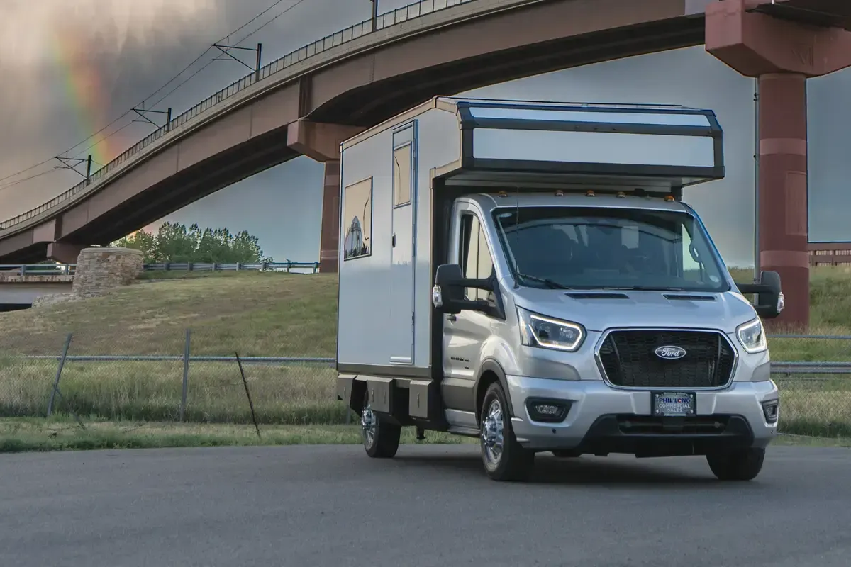 White Foundation Vehicles RampDeck body on a Ford Transit 156 driving on a highway beneath a dramatic storm sky with a rainbow