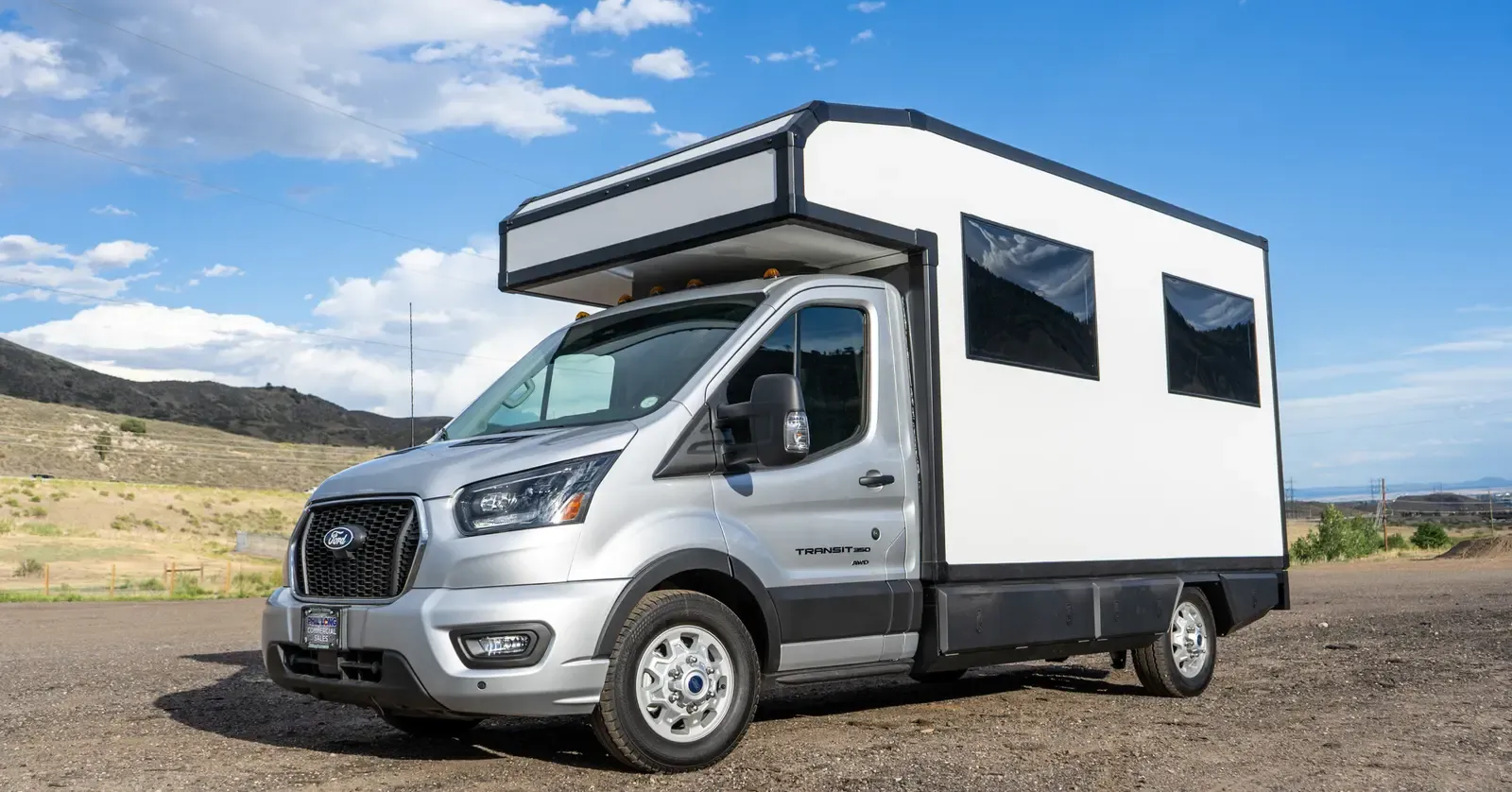 Silver Ford Transit 156 with a white composite RampDeck body by Foundation Vehicles, rear wall closed, front three-quarter view on a gravel clearing below the Colorado foothills